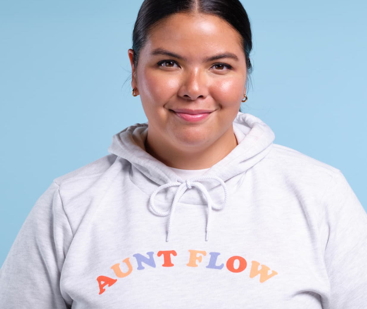 A woman with dark hair pulled back wears a light gray heather hoodie with 'AUNT FLOW' printed across the chest in orange and blue letters, set against a solid light blue background.