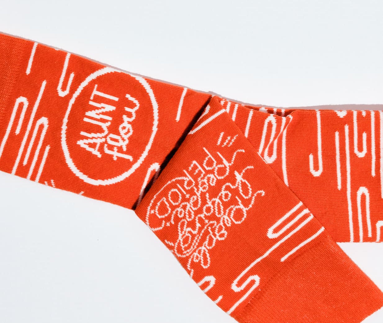 Bright orange knit scarf with white abstract line patterns and a circular logo reading 'AUNT FLOW' on a plain white background.
