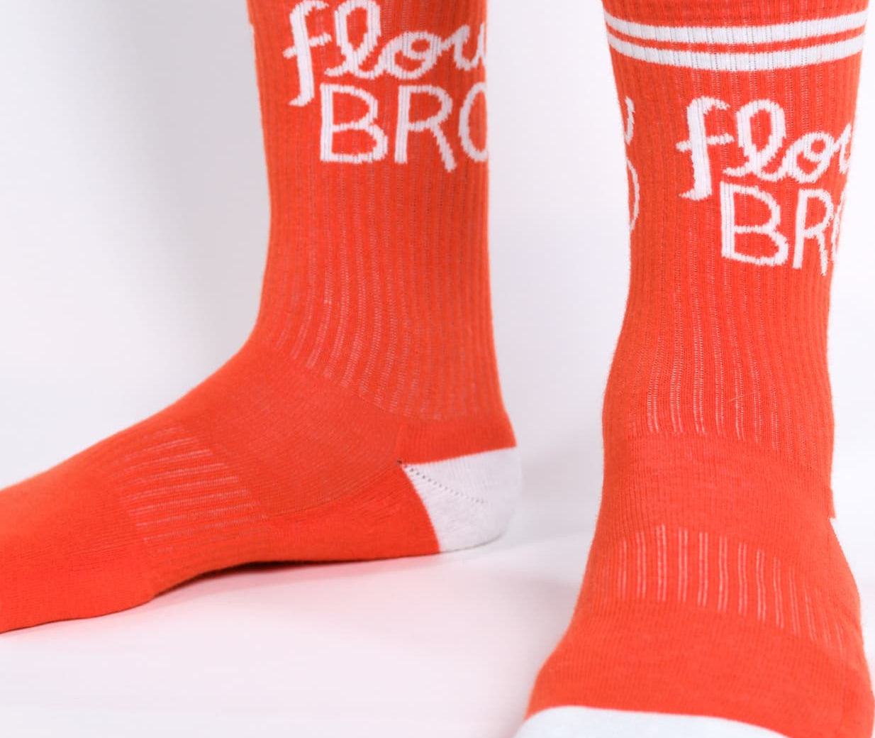 Bright orange crew socks worn on feet, featuring white stripes near the top and the words 'flow BRO' printed in white script and block lettering. The toes and heels are solid white.