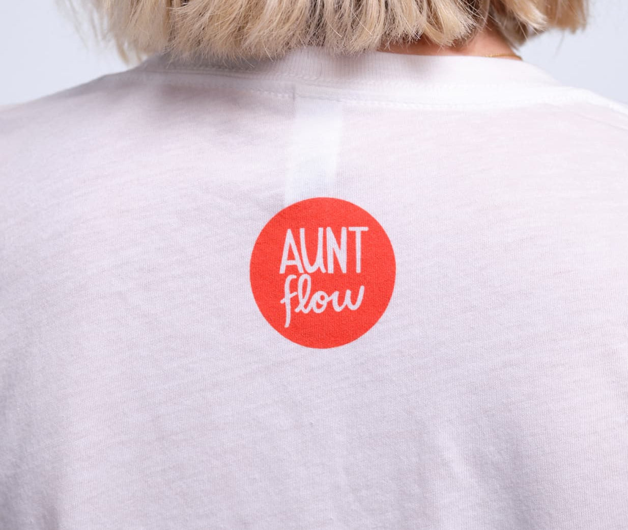 A close-up back view of a person with blonde hair wearing a white t-shirt with a red circular sticker that reads 'AUNT flow' in white text.