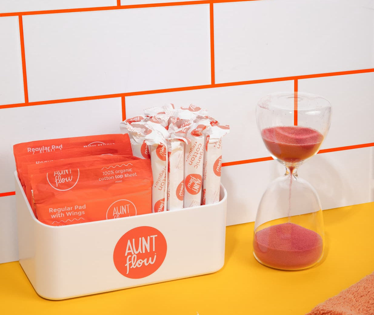 A white plastic display box with product divider, labeled with 'Aunt Flow', containing red period care pads and applicator tampons, placed on a yellow surface against a white tiled wall with orange grout. A glass hourglass with red sand is also visible to the side.